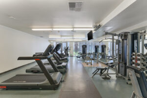 Interior Fitness Center, Tile Flooring, White wall, Mirrored wall, tv above treadmill station, multiple treadmills, stationary bikes, weightlifting equipment, exit to outside by windows.