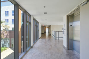 Interior Hallway, Floor to ceiling glass windows, elevator with hand sanitizer on the outside, tile flooring.