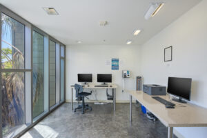 Interior Business center, 3 desktops, 1 printer, area fan, white walls, tile floor, floor to ceiling glass windows.
